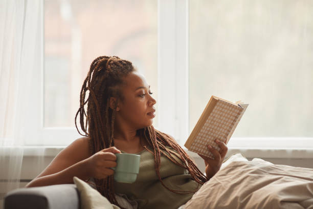 Black young woman lying on sofa drinking tea and reading an interesting book at home