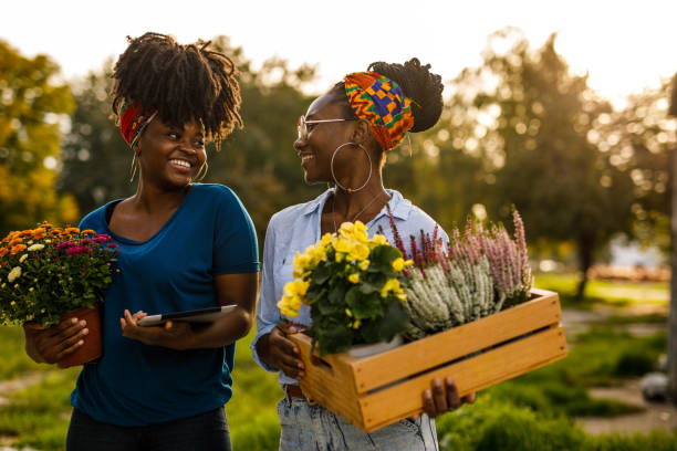 Portrait of two charming young Black florists standing on the city street, holding potted flowers, chatting casually.