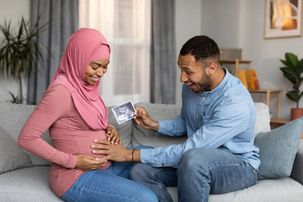 Excited Black Man Holding Baby Sonography Photo And Touching Pregnant Muslim Wife's Belly While They Relaxing Together On Couch At Home, Happy Islamic Spouses Enjoying Upcoming Parenthood